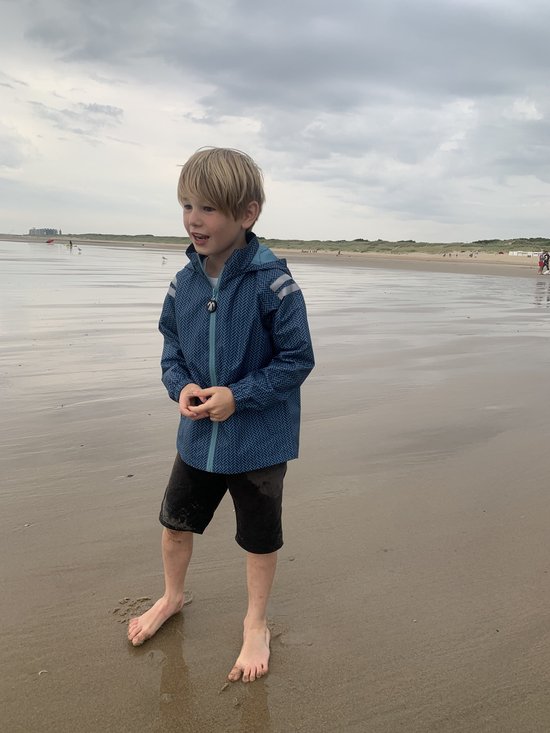 Jongen staand op het strand in een blauw regenpak met visgraatpatroon.