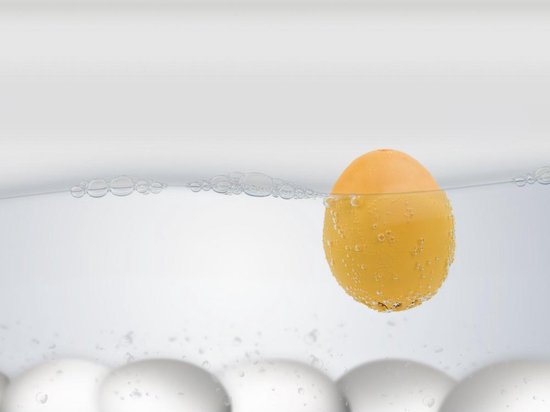 Gele eierwekker drijft in water met bubbels, witte eieren op de achtergrond.