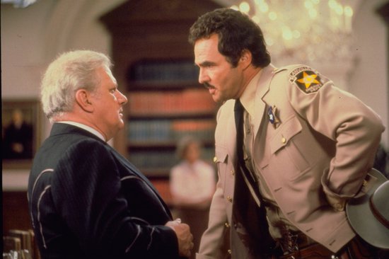 Twee mannen in gesprek: een in zwart pak, de ander in een beige sheriffuniform met pet.