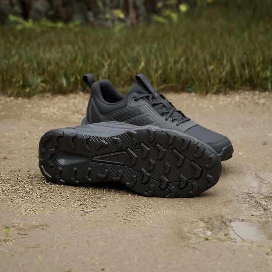 Zwarte Adidas Terrex trailrunningschoenen op een ondergrond van zand en gras, de ene schoen toont de zool.