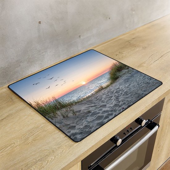 Een inductiebeschermer met een zonsondergang op een strand met duinen ligt op een houten aanrecht naast een inductiekookplaat.