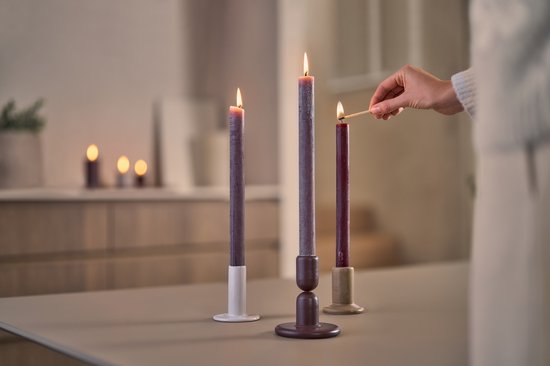 Hand steekt een paarse kaars aan in een bruine kandelaar. Twee andere paarse kaarsen staan ernaast.
