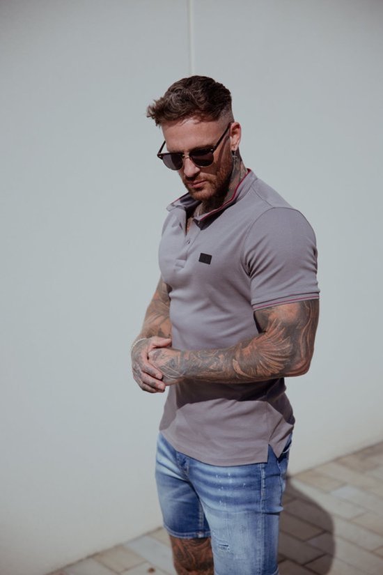 Man met tatoeages draagt een lichtgrijze polo met rode bies langs de kraag en mouwen. Hij draagt een zonnebril.