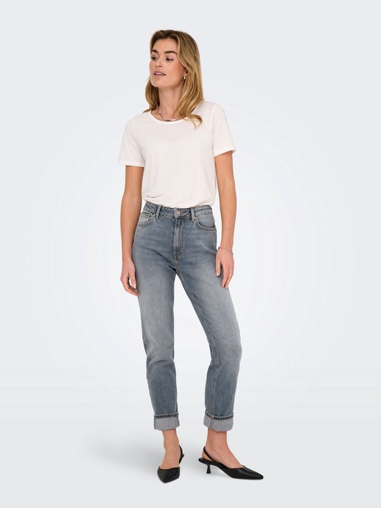 Vrouw in een witte T-shirt en grijze spijkerbroek met omgeslagen pijpen, staand.