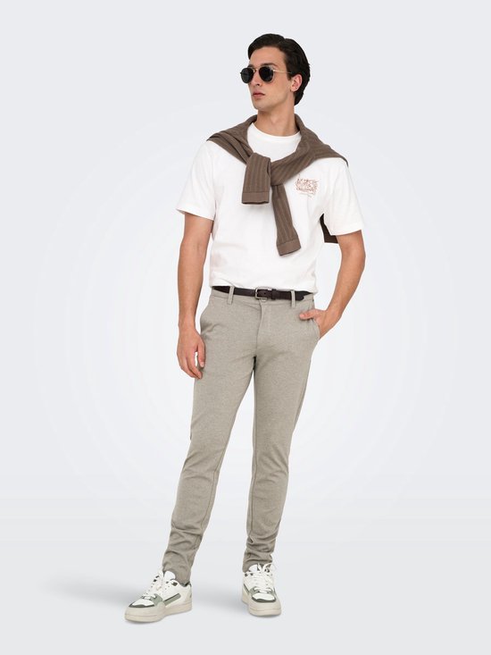 Man in een beige chino, wit T-shirt met print, bruine trui over de schouders, zonnebril en sneakers.