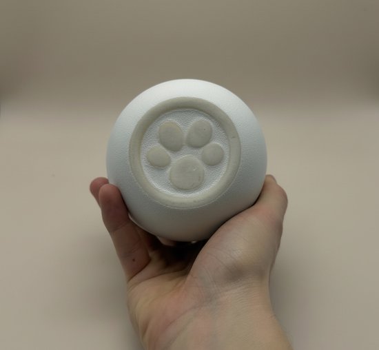 Een hand houdt de onderkant van een witte keramische urn vast. De bodem heeft een reliëf van een pootafdruk.
