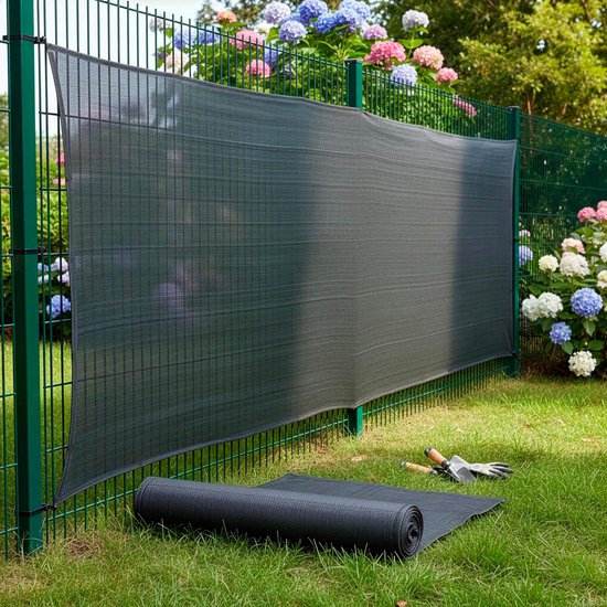 Een donkergrijs schaduwdoek is bevestigd aan een groene metalen hek, met bloeiende hortensia's erachter. Een opgerolde rol ligt op het gras.