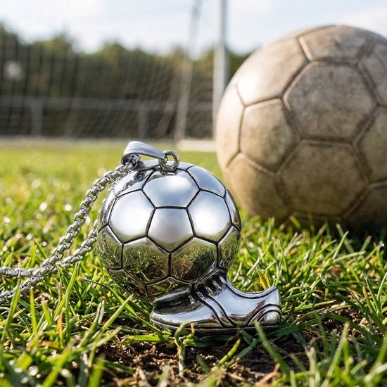 Gedetailleerde zilverkleurige voetbal- en schoenhanger aan een schakelketting op gras, met een voetbal op de achtergrond.