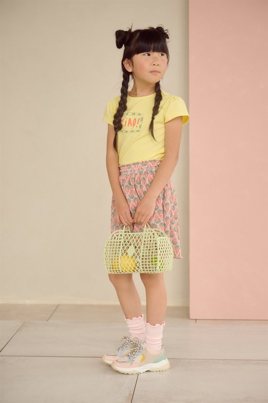 Meisje met zwarte staartjes draagt een gele "CIAO!" T-shirt en een lichtroze plooirok met hartjesprint.