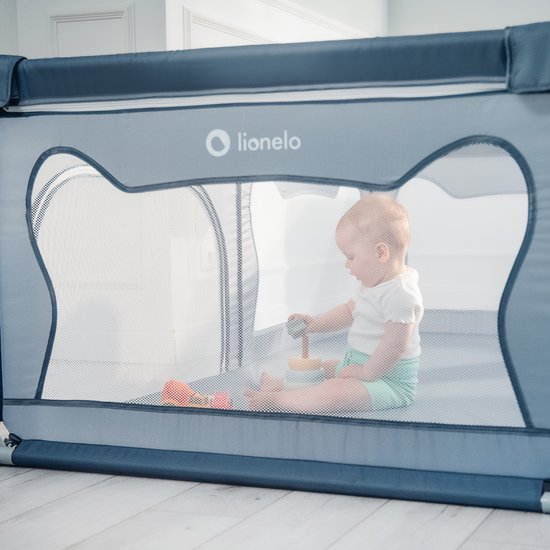 Baby in witte kleding speelt met houten speelgoed in een grijze box, gezien door gaas.