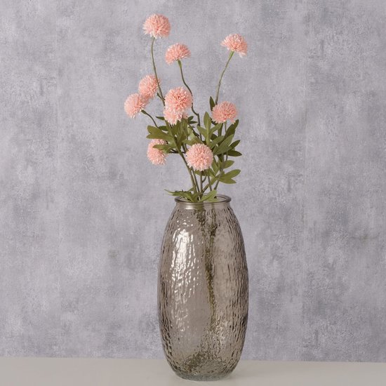 Hoge, bruin-beige glazen vaas met een ruw, bobbelig oppervlak, gevuld met lichtroze bloemen, tegen een grijze achtergrond.