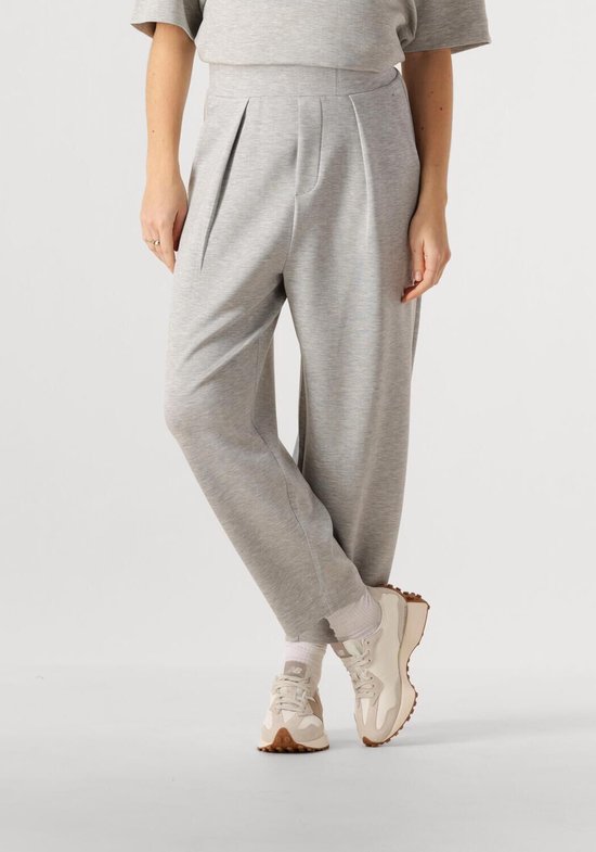 Model draagt een lichtgrijze broek, plooien aan de voorkant, witte sokken en sneakers.