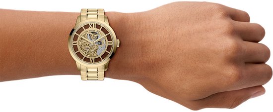 Pols met een goudkleurig Fossil skeleton horloge met metalen band, op de achtergrond is een deel van de hand te zien.