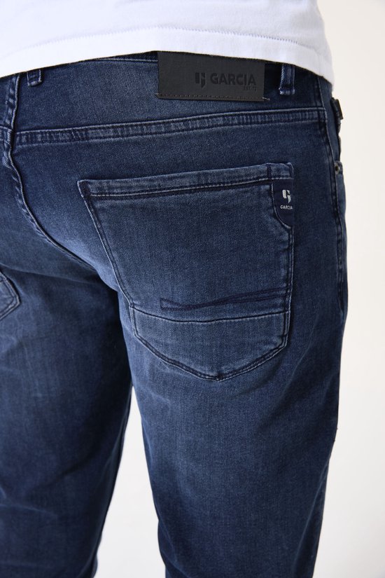 Achterkant van een donkerblauwe jeans met Garcia logo op de zwarte achterzak patch.