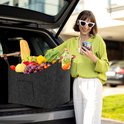 Lachende vrouw met zonnebril houdt een smartphone vast naast een donkergrijze vilten mand gevuld met boodschappen.