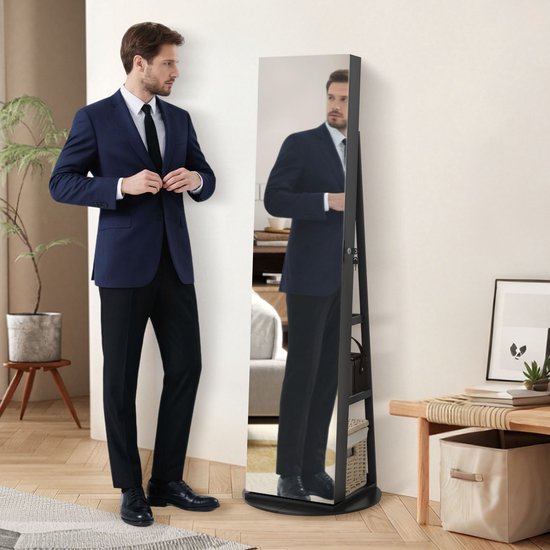 Man in een blauw pak staat naast een zwarte staande spiegelkast, die een slaapkamer reflecteert.