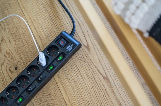Zwarte stekkerdoos met 6 stopcontacten en individuele schakelaars, een witte USB-kabel aangesloten.