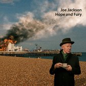 Joe Jackson op een kiezelstrand, met op de achtergrond een brandende pier. Joe Jackson, Hope and Fury.