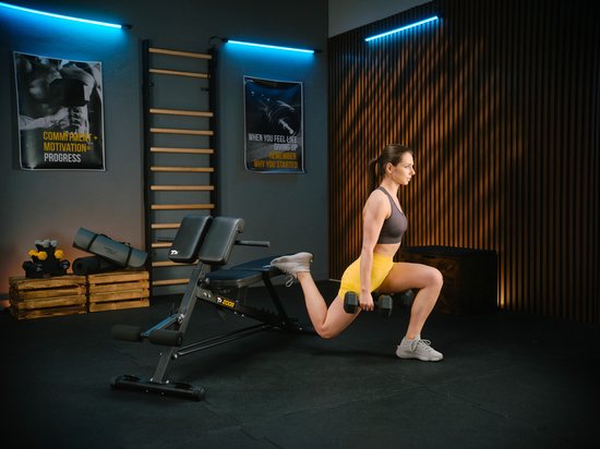 Vrouw met een sporttop en gele shorts doet een lunges met dumbbells op een zwarte fitnessbank.