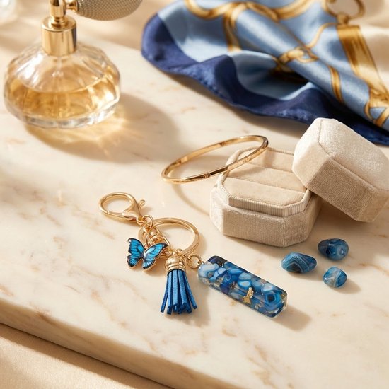 Gouden sleutelhanger met blauwe vlinder, blauw kwastje en rechthoekige hanger ligt naast een parfumflesje en blauwe sjaal.