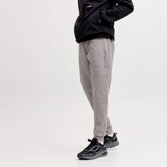 Man met grijze joggingbroek, zwarte jas en zwarte sneakers, staand op witte achtergrond.