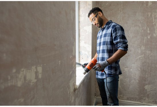 Man met veiligheidsbril, een blauw geruit shirt en handschoenen gebruikt een oranje Black+Decker verfbrander bij een raam.