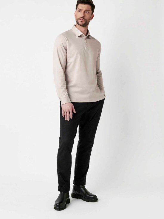 Man in beige longsleeve poloshirt en zwarte broek, staand.