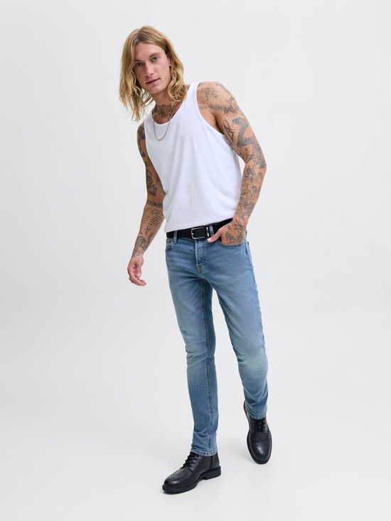 Man met blonde haren, draagt een witte tanktop en blauwe jeans met lichte wassing, zwarte riem en zwarte schoenen.