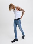 Man met blond haar en tatoeages draagt een lichtblauwe skinny jeans, witte tanktop en zwarte schoenen.