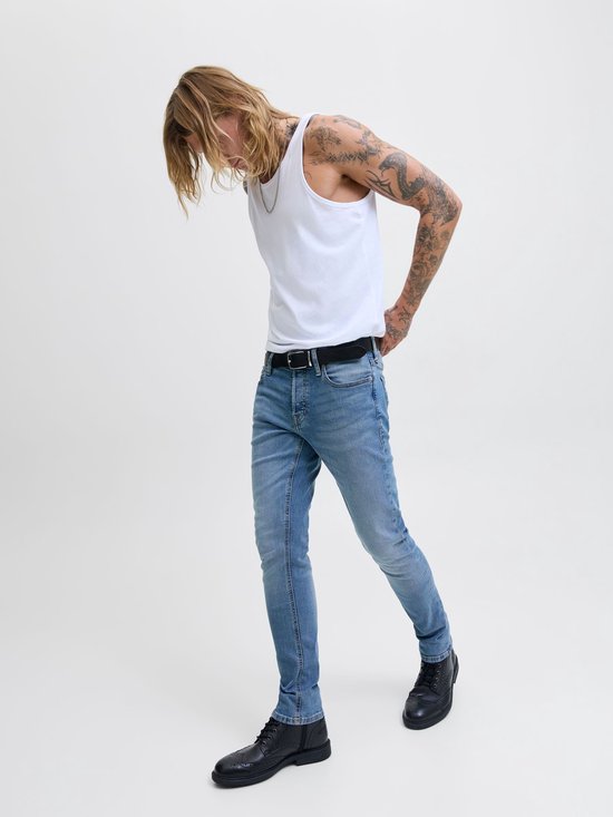 Man met blond haar en tatoeages draagt een lichtblauwe skinny jeans, witte tanktop en zwarte schoenen.