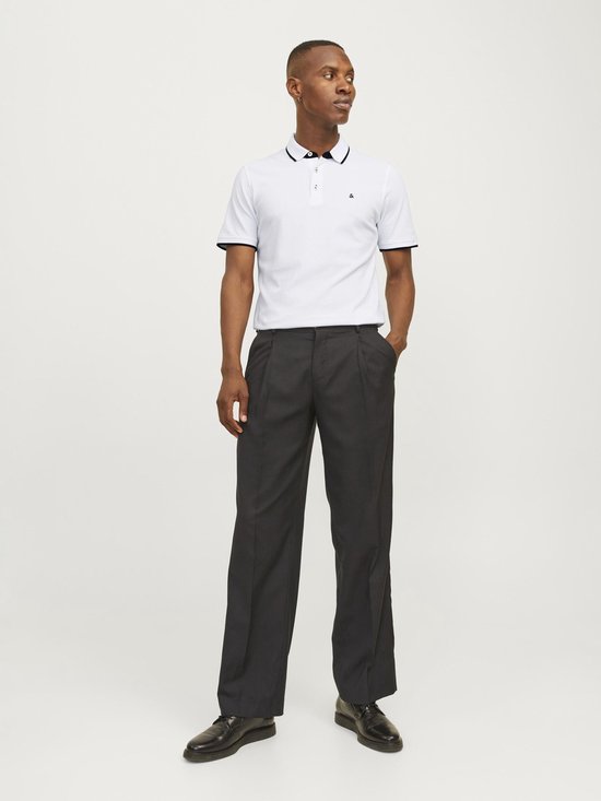 Man staat, draagt witte poloshirt met zwarte kraag en biezen, en donkergrijze pantalon.