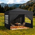Zwarte partytent in een veld met bergen op de achtergrond, met een opgevouwen tafel en twee stoelen binnen.