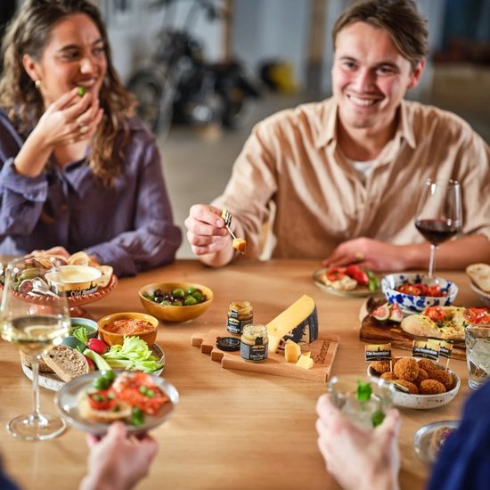 Een houten tafel met mensen die Old Amsterdam kaas en hapjes delen, met kaasblokjes en mini-chutneys.