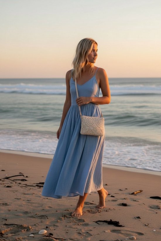 Blauw maxi-jurk met spaghettibandjes, gedragen door een blondine op het strand met een gehaakt tasje.
