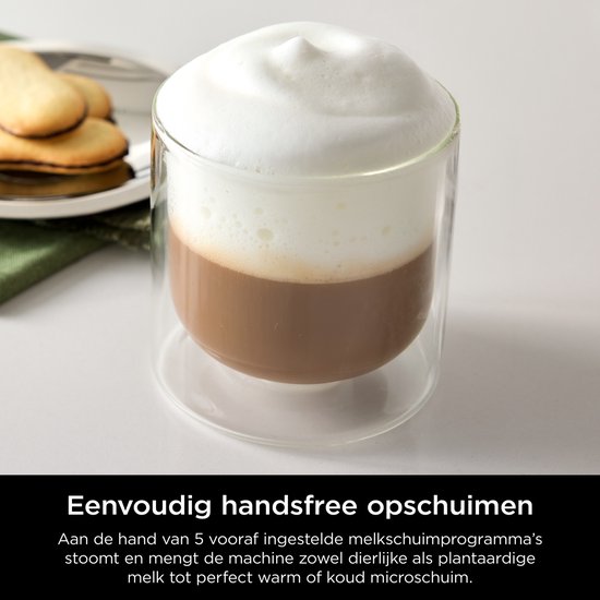 Glazen mok met warme chocolademelk en een dikke laag melkschuim, naast koekjes.