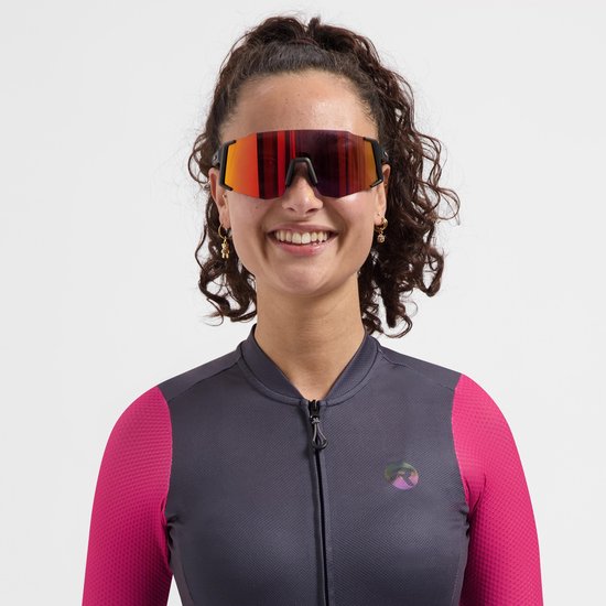 Vrouw met krullend haar draagt een zwarte Rogelli Sabre sportbril met gespiegelde rode lenzen en een grijs-roze fietsjack.