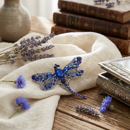 Blauwe libelle broche met zilverkleurig metaal en fonkelende facet geslepen stenen, op een beige doek met lavendel.