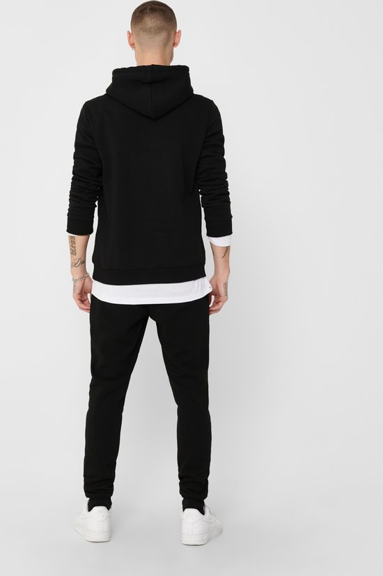 Zijaanzicht van man met tatoeages die een zwarte hoodie met capuchon draagt met een witte longsleeve eronder.