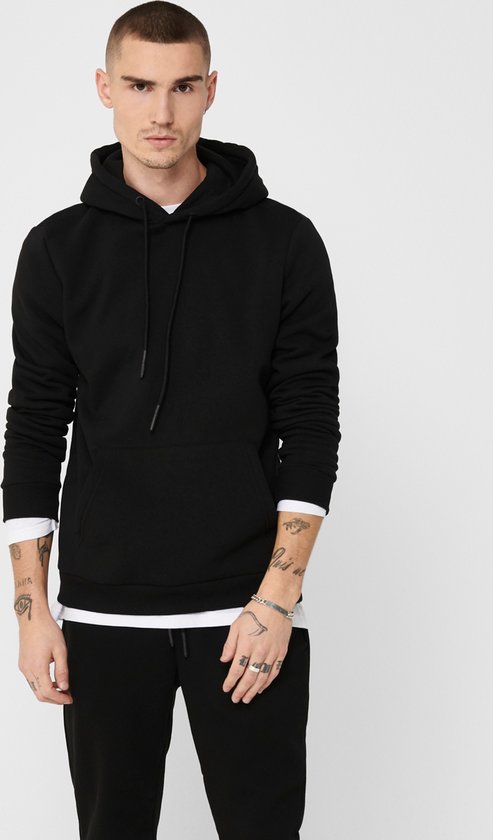 Man met kort bruin haar draagt een zwarte hoodie over een wit T-shirt, met spijkerbroek.