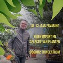 Man in grijze jas, glimlachend, staand in een tuincentrum met veel planten.