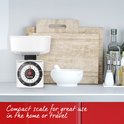 Compacte witte keukenweegschaal met zwarte wijzerplaat en rode wijzer, op een aanrecht.