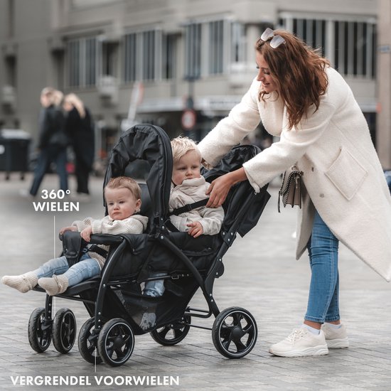 Zwarte dubbele buggy met twee kinderen erin, een vrouw duwt de buggy.