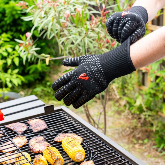 Twee handen met zwarte, hittebestendige handschoenen met siliconen noppen boven een barbecue.