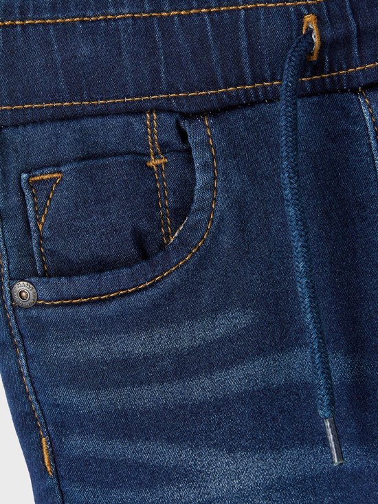 Detail van donkerblauwe spijkerbroek met stonewashed effect. Oranje stiksels, trekkoord en knoop aan de voorzijde.