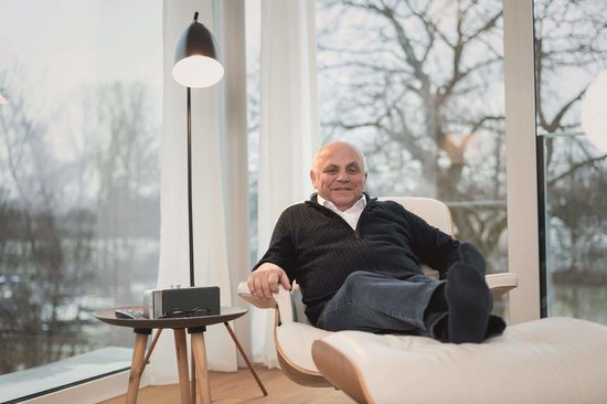 Een zittende man met een afstandsbediening op een stoel in een woonkamer. Een luidspreker met zilverkleurig handvat staat op een bijzettafel.