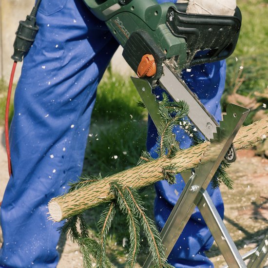 Een persoon in blauwe overall zaagt met een elektrische kettingzaag een dennengroeisel door op een zaagbok.
