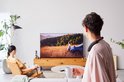 Man met nekband speaker, drinkt koffie en kijkt tv.