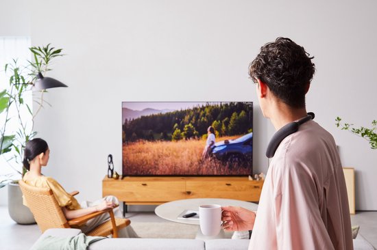 Man met nekband speaker, drinkt koffie en kijkt tv.