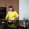 Een vrouw in een gele trui bakt een ei in een koekenpan in een keuken.