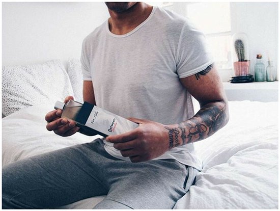 Man met tatoeages zit op een bed, vasthoudend aan een flacon 'Van Gils Between Sheets'.